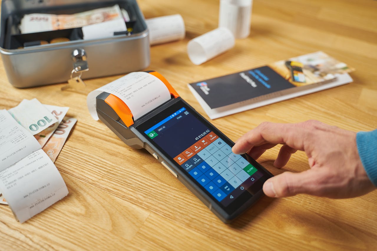 about-us Person using a digital payment terminal on a wooden desk with cash and receipts visible.